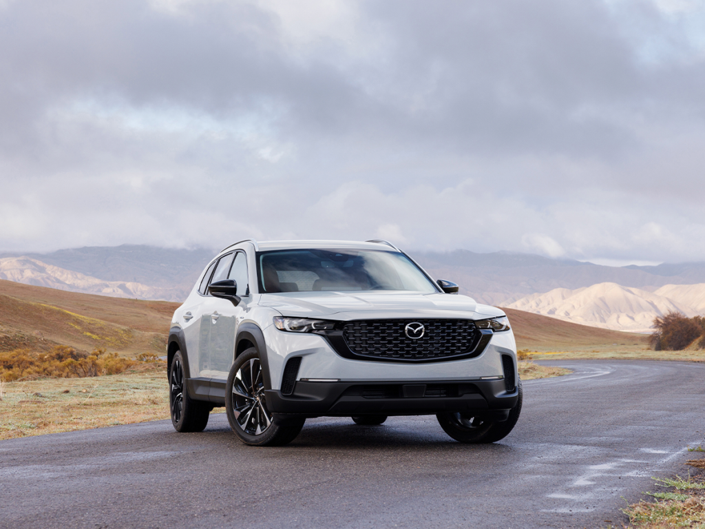 Exterior front view of a white Mazda CX-50 parked on a winding road with mountains in the background under a cloudy sky.
