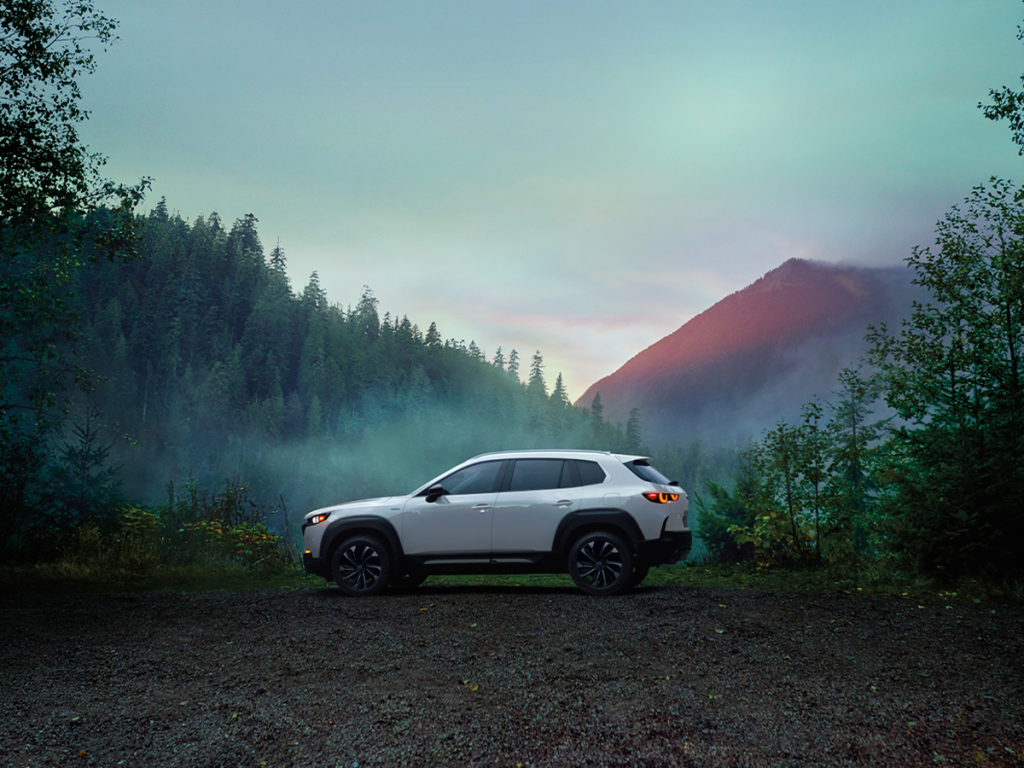 Side view of a white Mazda CX-50 parked in a scenic forest area, surrounded by misty trees and a mountain in the distance.