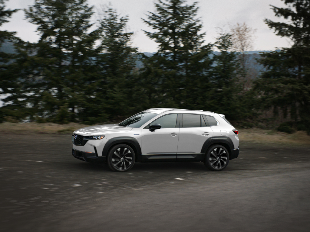 Side profile of a white Mazda CX-50 driving on a tree-lined road, capturing the dynamic and sporty design of the vehicle in motion.