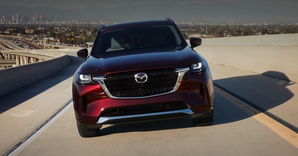 Front view of a red Mazda CX-90 SUV driving on an open highway with a cityscape in the background."